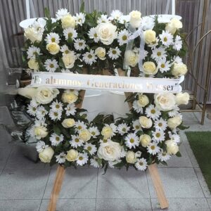 Corona fúnebre blanca con rosas, margaritas y cinta de condolencias sobre atril de madera para funerales en Concepción.