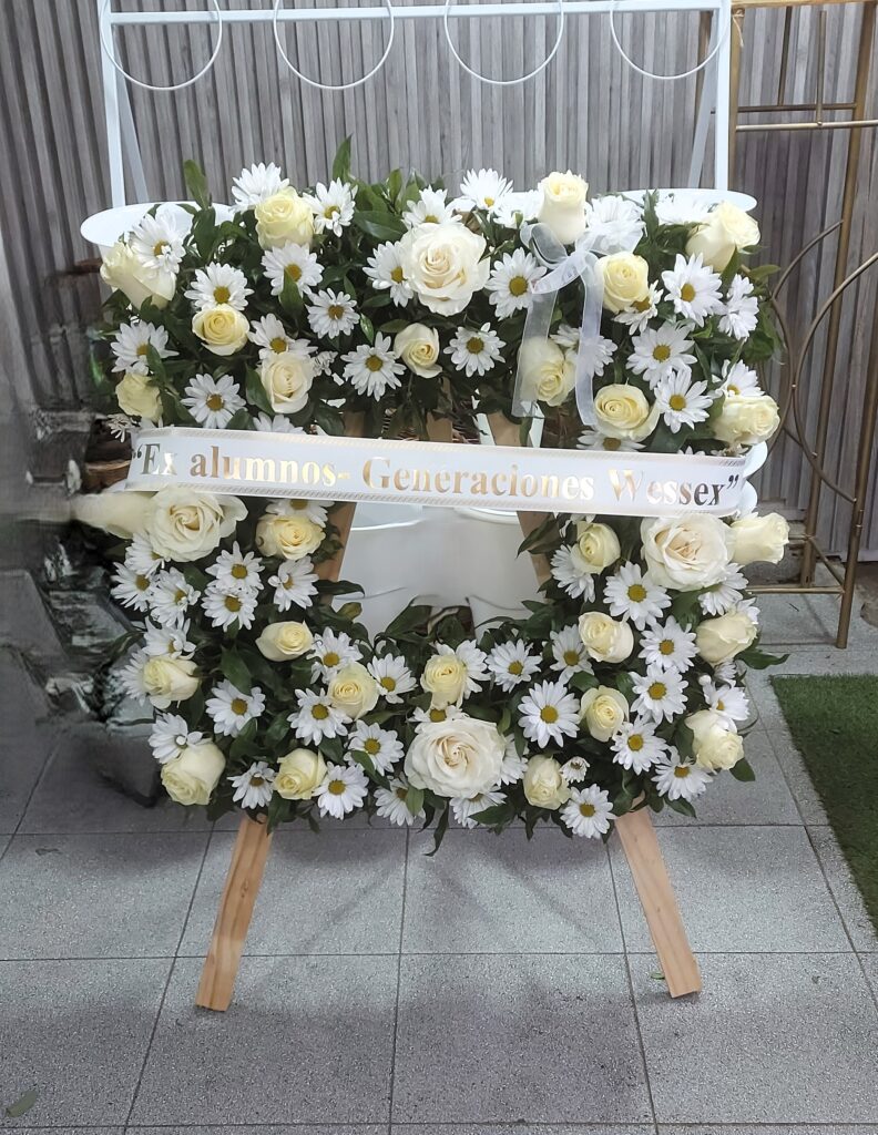 Corona fúnebre blanca con rosas, margaritas y cinta de condolencias sobre atril de madera para funerales en Concepción.